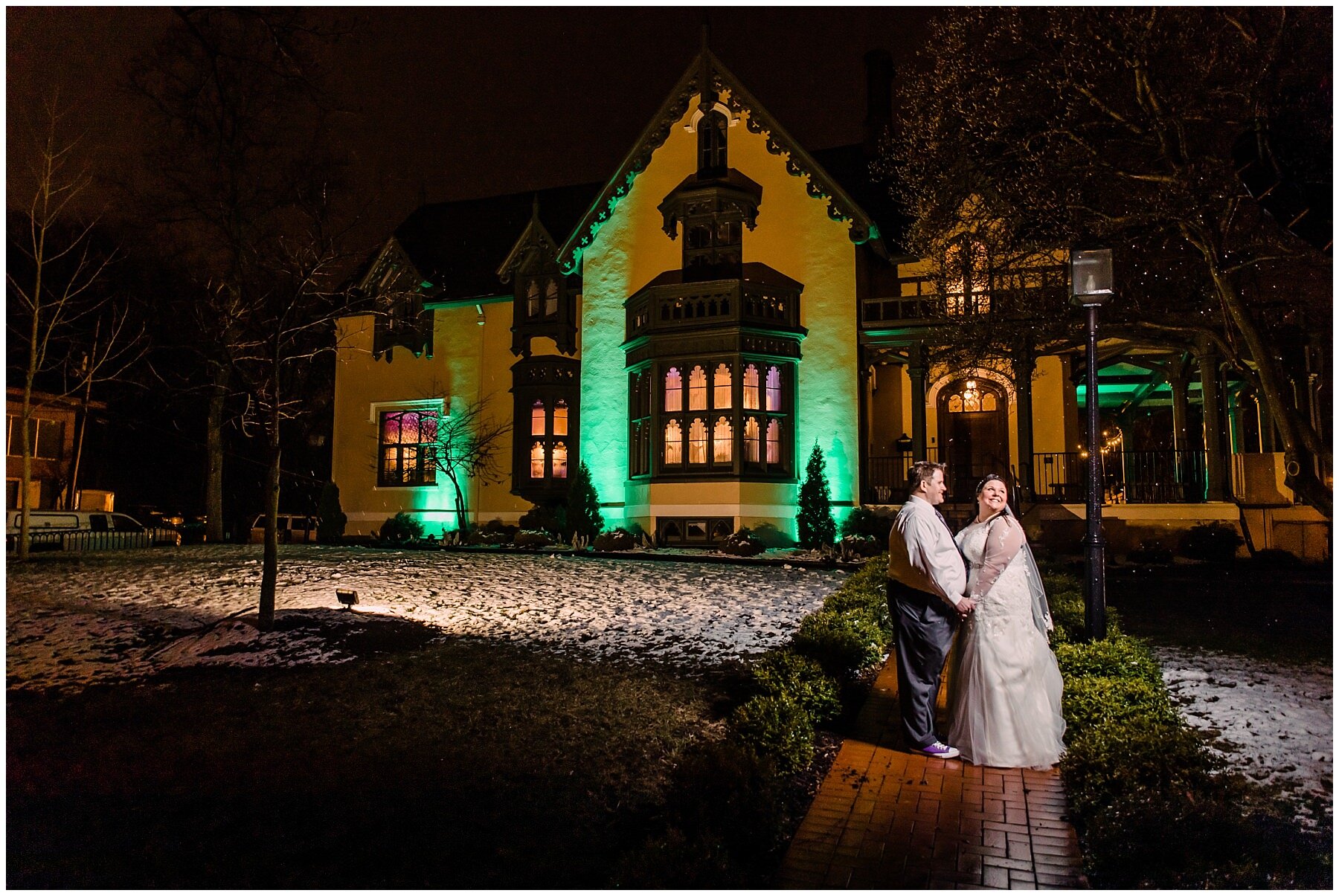 bride and groom standing in front of a historical mansion in lafayette indiana at night looking off at a light with snow around them