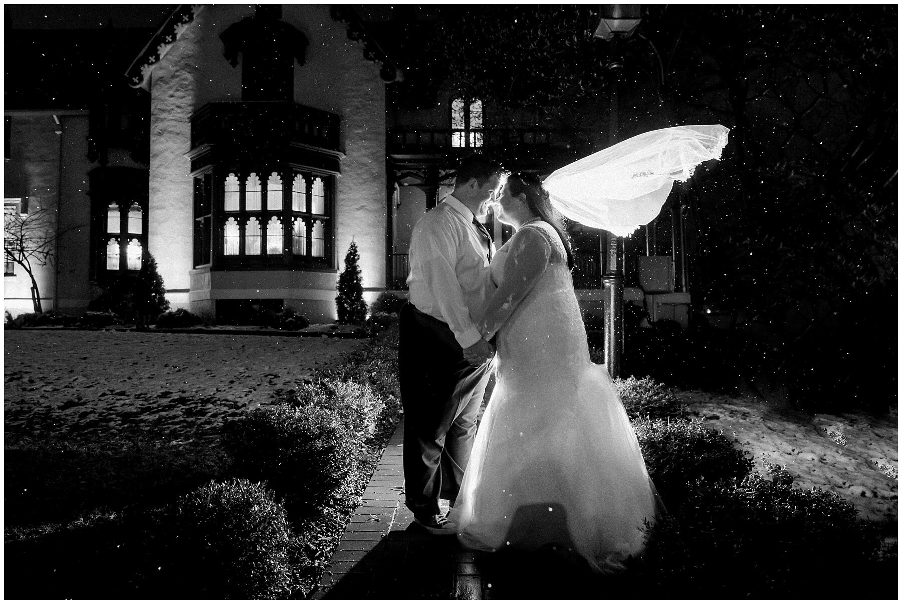 bride and groom standing in front of a mansion with snow falling around them at night veil flying back