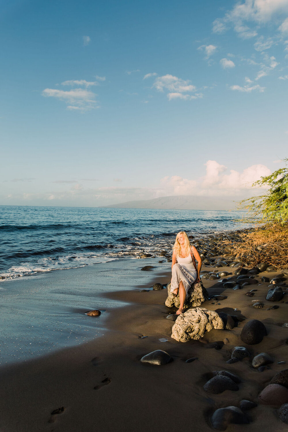 blonde woman sitting on a beach on a rock facing the sunrise in hawaii maui smiling