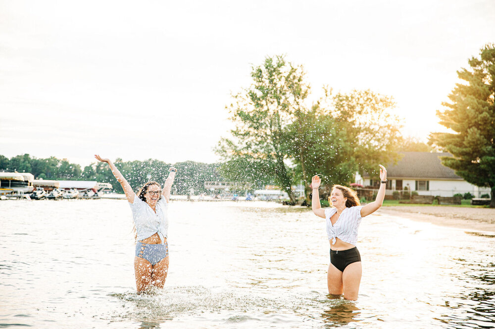 lesbian couple engagement session playing in the water splashing in the sunset in swimsuits