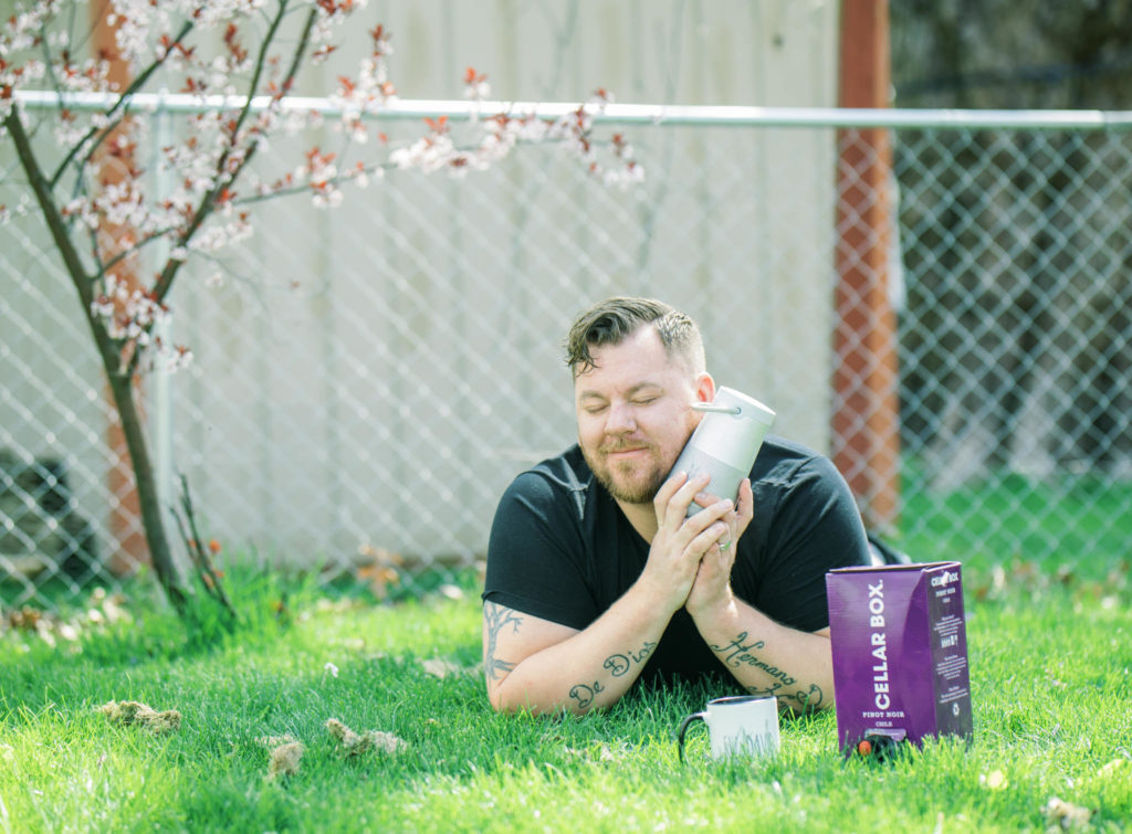 man listening to music with a box of wine and a coffee mug in the grass on a bose revolve speaker thinking about how to look and feel beautiful during your engagement photos
