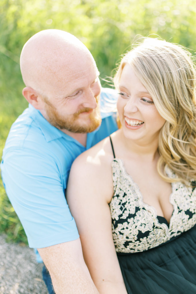 happy couple sitting on the ground laughing and smiling with golden light while looking and feeling beautiful during their engagement photos